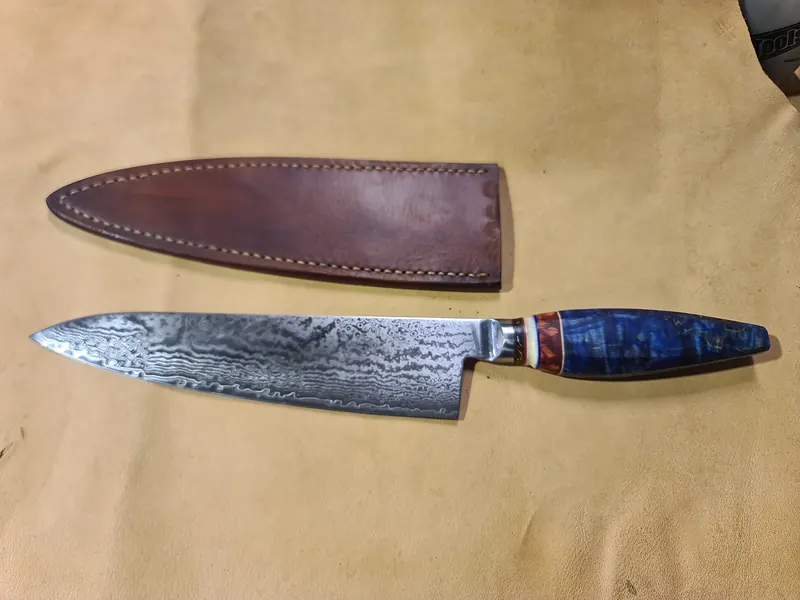 Blue-handled chef’s knife with a patterned blade below its stitched brown leather sheath